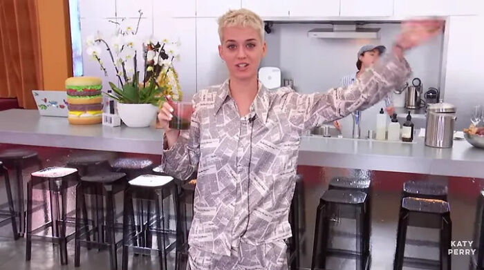 A person in patterned pajamas holding a drink, waving in a modern kitchen setting with stools and plants, pop culture cringe moment.