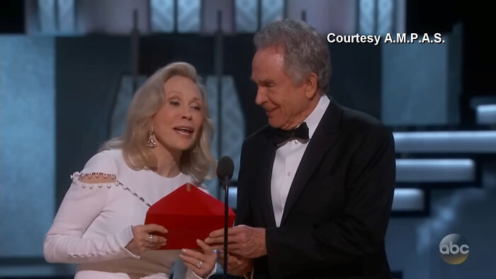An elderly woman and man in formal attire holding a red envelope on stage during a pop culture moment that made people cringe hard.