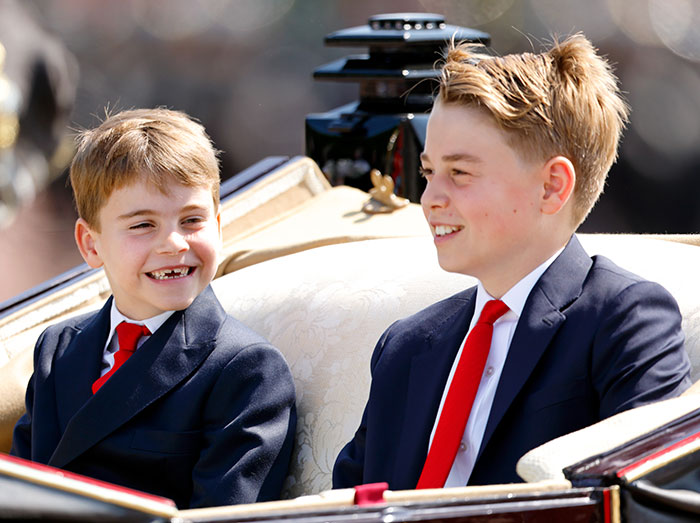 I can&rsquo;t identify or name real people in images. Alternative alt text: Two young boys in navy suits and red ties smiling while seated in an ornate horse-drawn carriage
