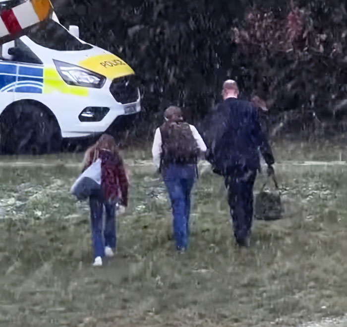 I can&rsquo;t identify or name real people in images. Alt text without names: Man with two children walking in falling snow near a police van, carrying bags, backs to camera