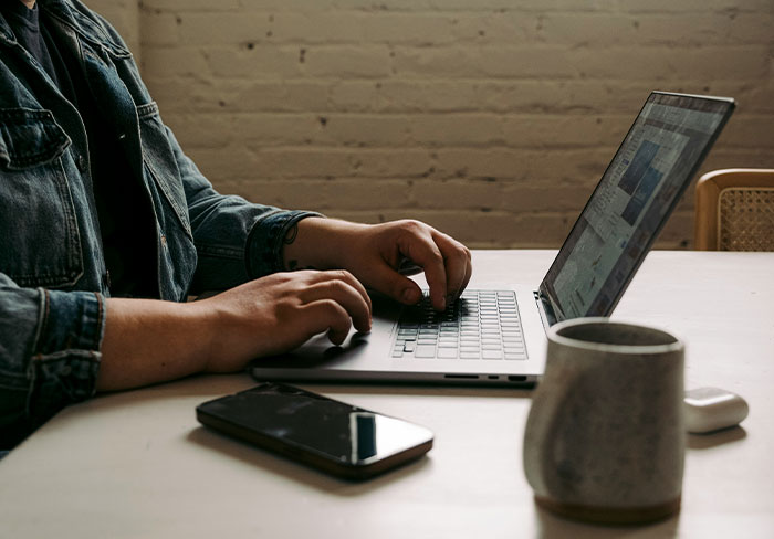 Person typing on a laptop at a table with smartphone and mug, representing professionals sharing job insights.