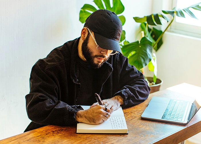 Man wearing a cap and glasses writing notes at a desk with a laptop, illustrating professionals sharing job insights.