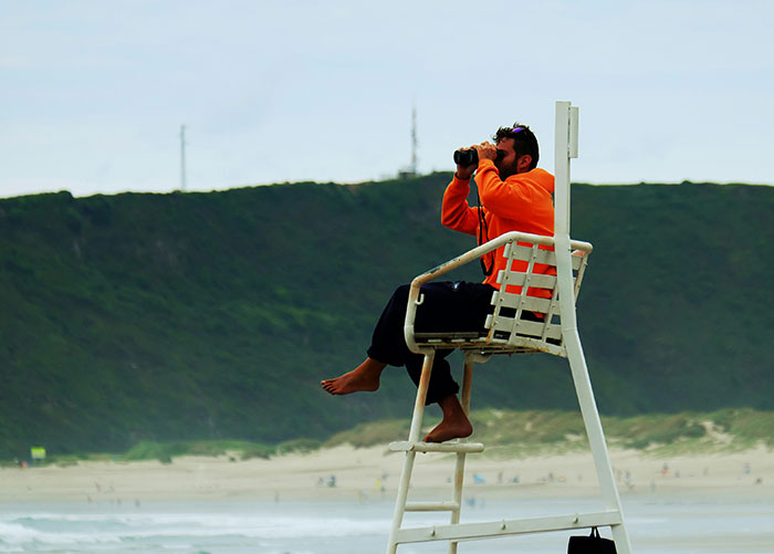 Lifeguard in an orange jacket using binoculars, highlighting insights from professionals about their jobs to the public.