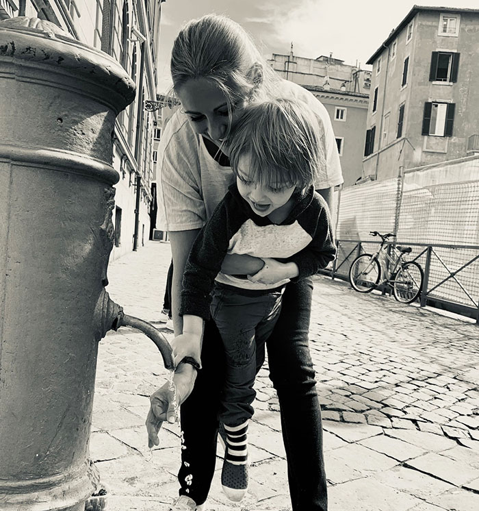 Woman lifting child to wash hands at outdoor water fountain on cobblestone street in urban area.