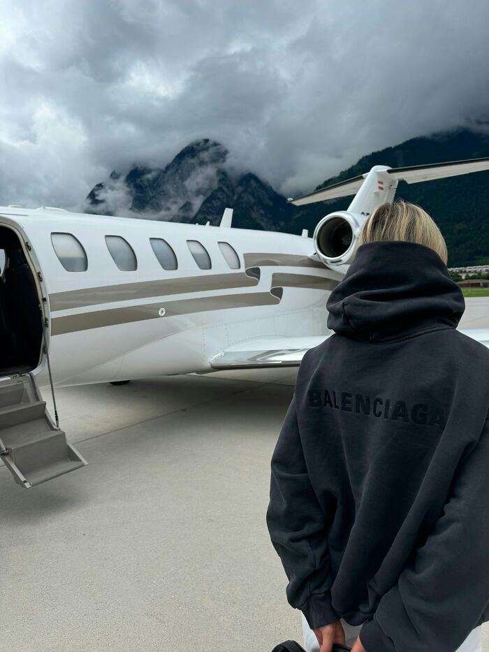 Person wearing Balenciaga hoodie standing near a private jet with mountains and cloudy sky in background showing rich people lifestyle