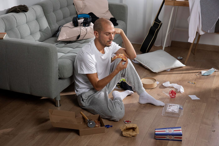 Man-child hubby sitting on messy floor eating pizza, surrounded by clutter in living room, reflecting working wife challenges. - 9