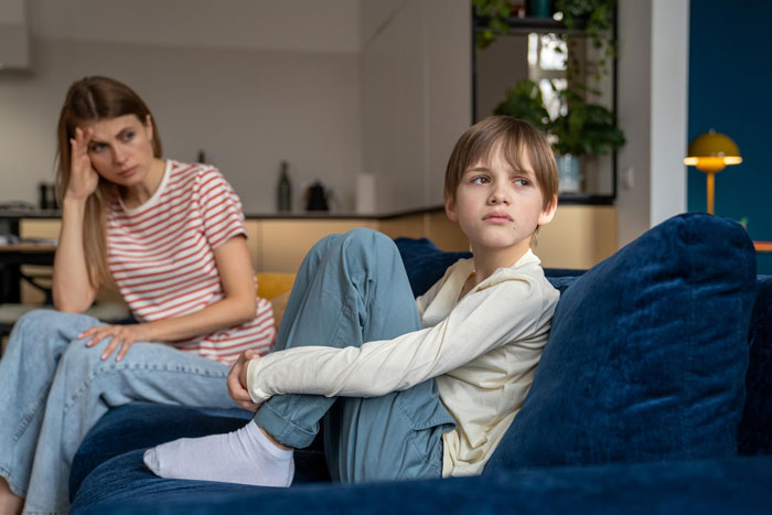 Sad autistic kid sitting on couch looking away while worried mom watches him upset in modern living room.