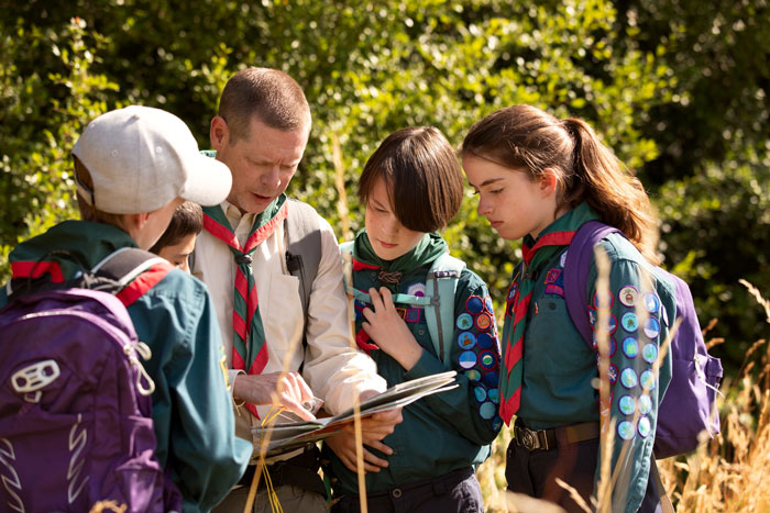 Scout leader showing a map to kids wearing uniforms and backpacks during an outdoor activity in a forest setting.