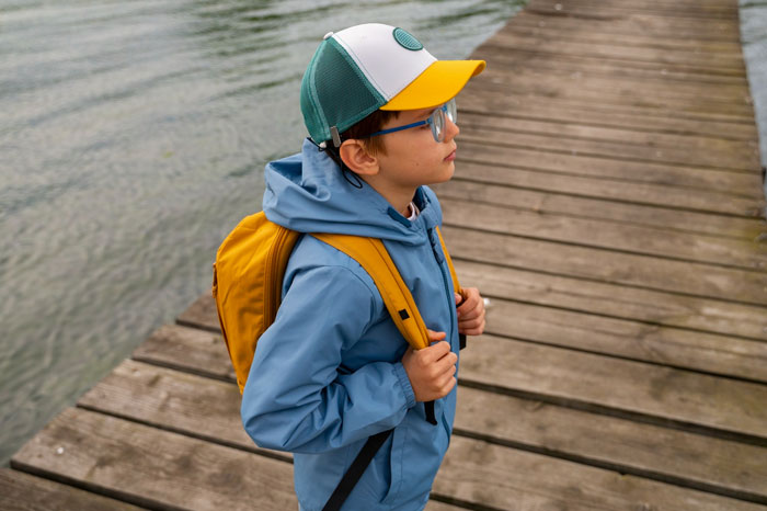 Young autistic kid wearing a cap and backpack, standing alone on a wooden dock near water, looking thoughtful.