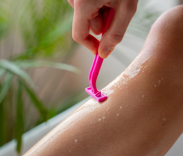Hand holding pink razor shaving wet leg emphasizing the topic of refuse shaving legs work and personal care choices.