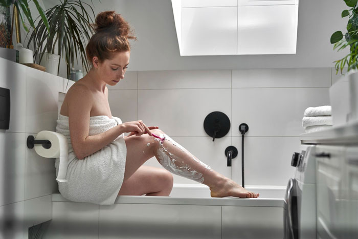 Young woman wrapped in a towel shaving her legs in a modern bathroom, addressing personal hygiene and grooming.