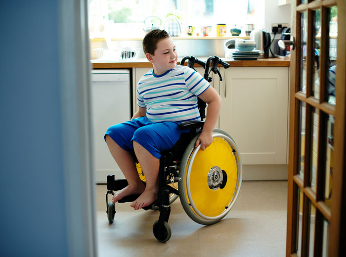 Boy with autism sitting in a wheelchair in a kitchen, highlighting challenges of caregiving and family responsibilities.
