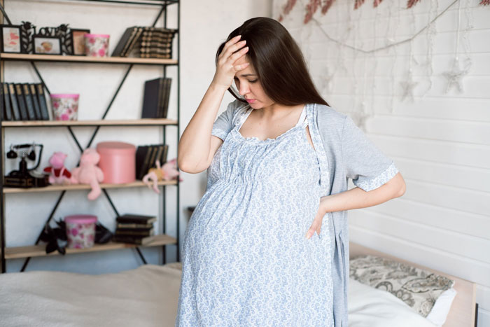 Pregnant woman looking distressed at home, holding head and standing by bed in a softly lit room with shelves.