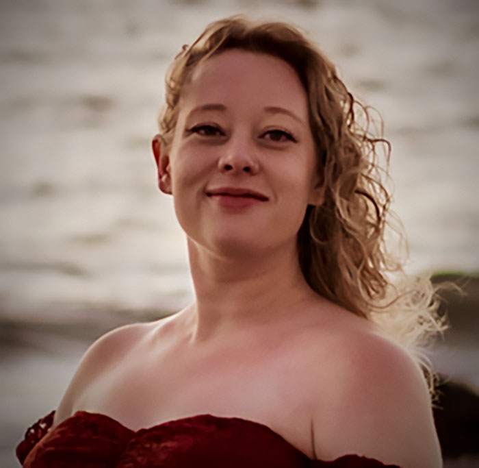 Young woman with curly blonde hair wearing a red off-shoulder dress near water, representing billionaire donation controversy.