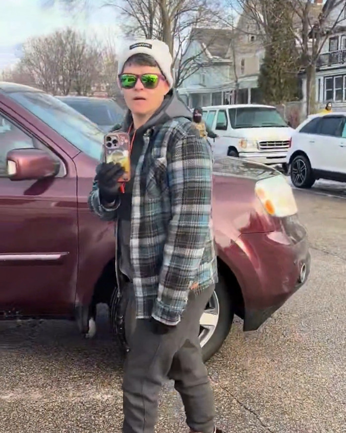 Person wearing green sunglasses and a hat, holding a smartphone near parked vehicles in an urban neighborhood scene.