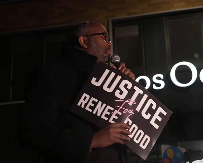 Man holding a Justice for Ren&eacute;e Good sign speaking into a microphone during a public gathering about fatal shooting context.