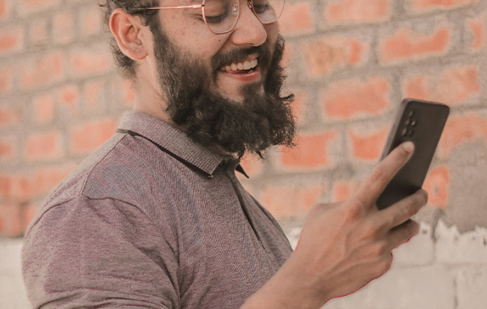 Man with glasses and beard smiling while looking at his phone, enjoying a moment of glorious revenge and the last laugh.