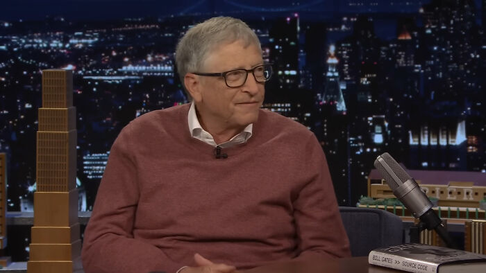 Man in glasses and maroon sweater discussing rich people norms on a late night talk show set with city lights background.