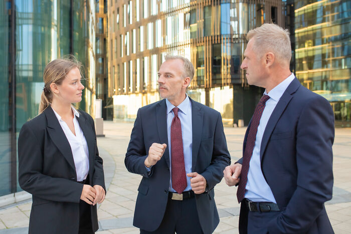 Three rich people in suits discussing business norms outdoors near modern glass buildings in a financial district setting