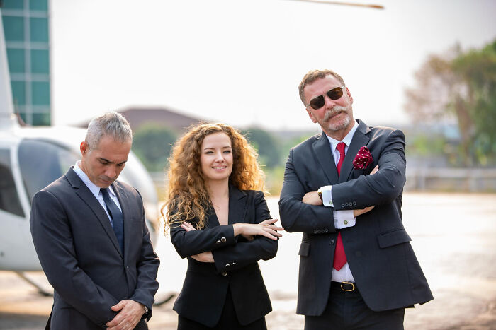 Three wealthy people dressed in business attire standing confidently near a private helicopter outdoors.