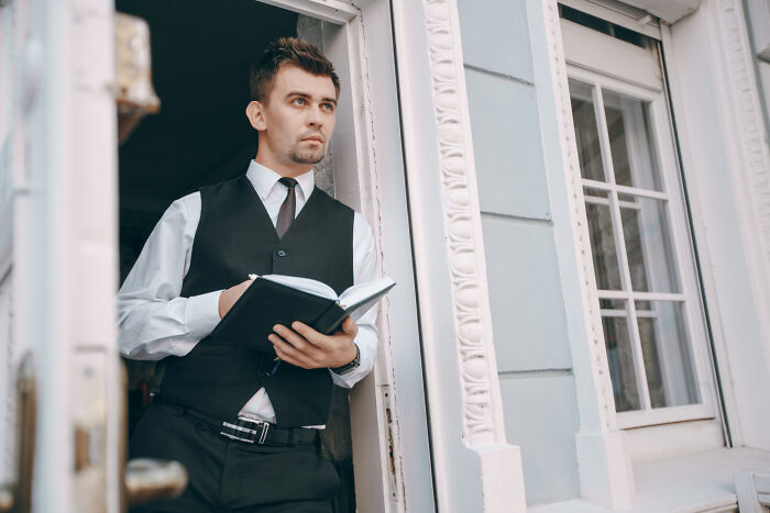 Man in a vest holding a notebook, standing in a doorway near a window, illustrating rich people norms insights.