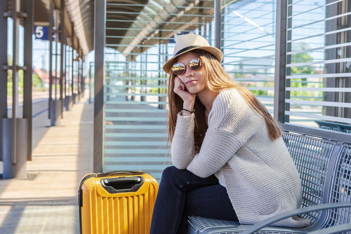 Young woman wearing sunglasses and a hat sitting with yellow suitcase at station, hinting roommate boyfriend vacation drama.