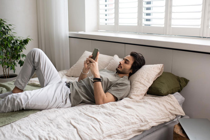 Young man lying on bed using smartphone, appearing relaxed and thoughtful in a softly lit bedroom setting.