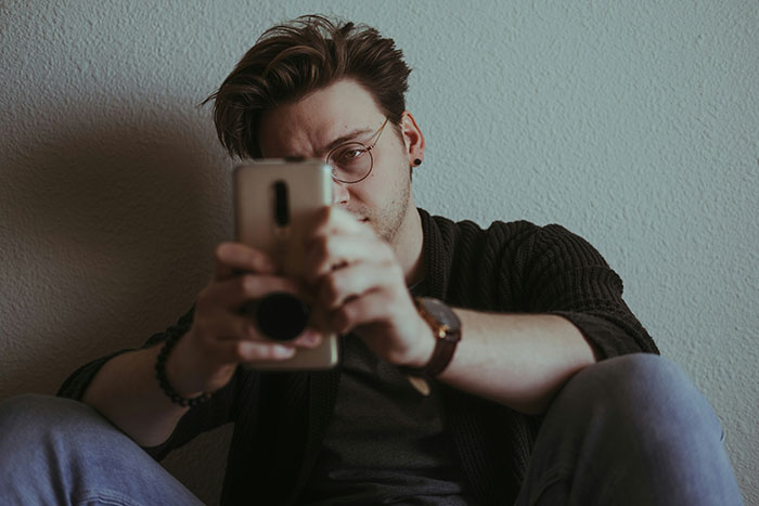 Young man with glasses sitting against a wall, holding phone, reflecting on superhero topics and grief-related emotions.