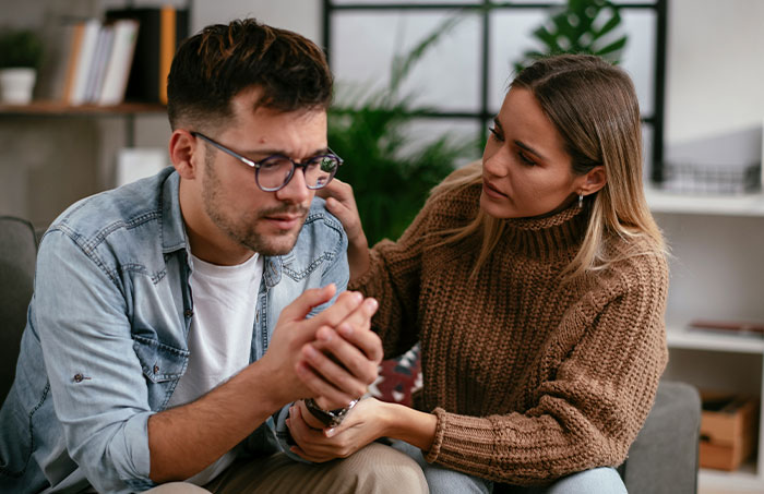 A woman comforting a distressed man in a living room, illustrating the impact of ruining family relationship lying MIL issues.