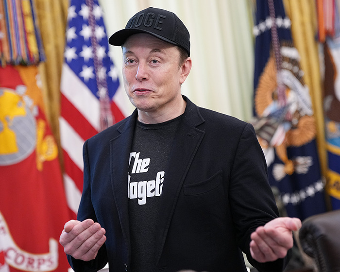 Elon Musk wearing a black cap and blazer, gesturing during a discussion with flags in the background.