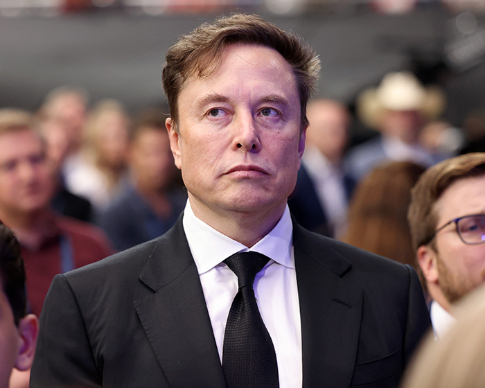 Elon Musk in a black suit and tie at an event, showing a serious expression amid a blurred crowd background.