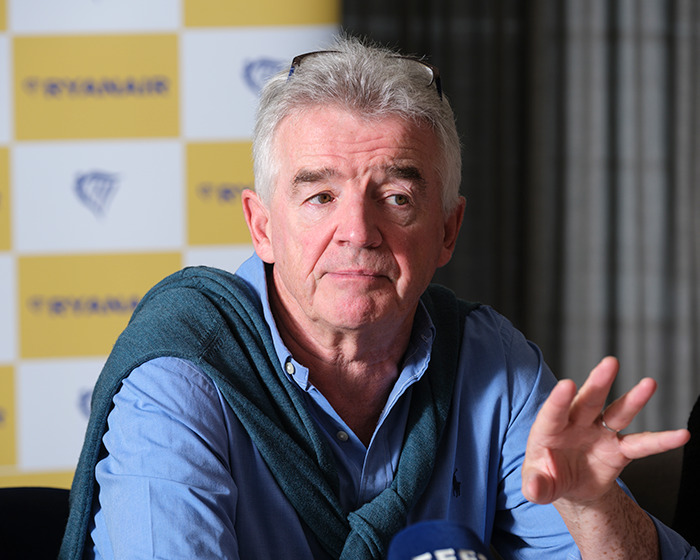 Man speaking at Ryanair press event, wearing blue shirt and sweater, with Ryanair logo backdrop, discussing airline response.