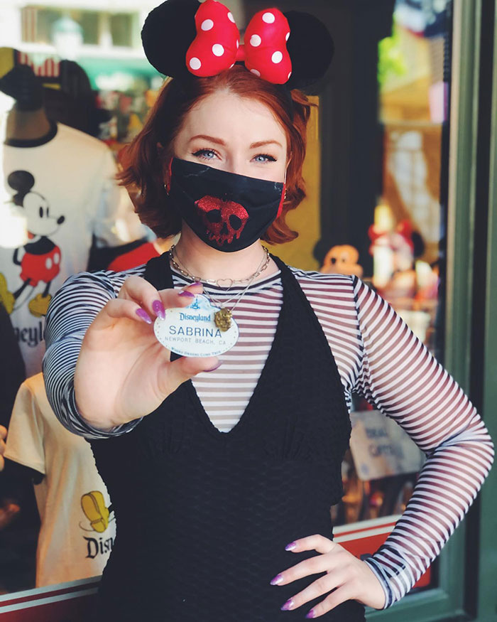 Woman in Minnie ears and skull mask holding Disneyland name tag revealing viral evil queen identity after getting fired.