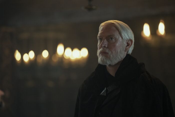 Actor portraying a Targaryen in A Knight of the Seven Kingdoms with white hair and beard in a dimly lit medieval setting.