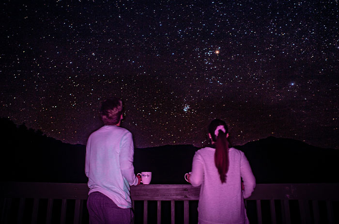 Two people on a deck stargazing with mugs under a vivid starry sky, saw images on Google