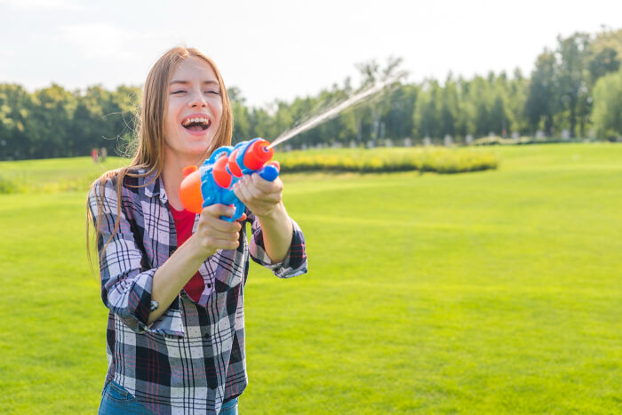 Teenage girl smiling outdoors, playfully squirting water with a toy gun, capturing a lighthearted high school bully moment.