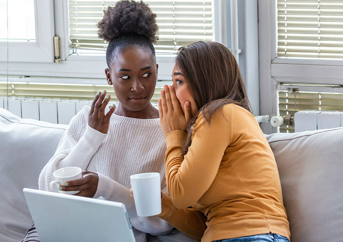 Two women sharing secrets on a couch with coffee cups, illustrating anonymous people exposing explosive secrets.