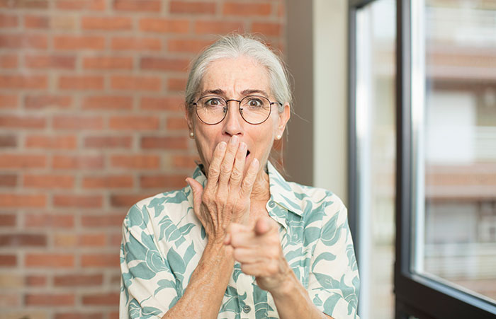 Older woman in glasses and patterned shirt looking shocked and pointing, representing wife done with hubs&rsquo; relatives living with them.