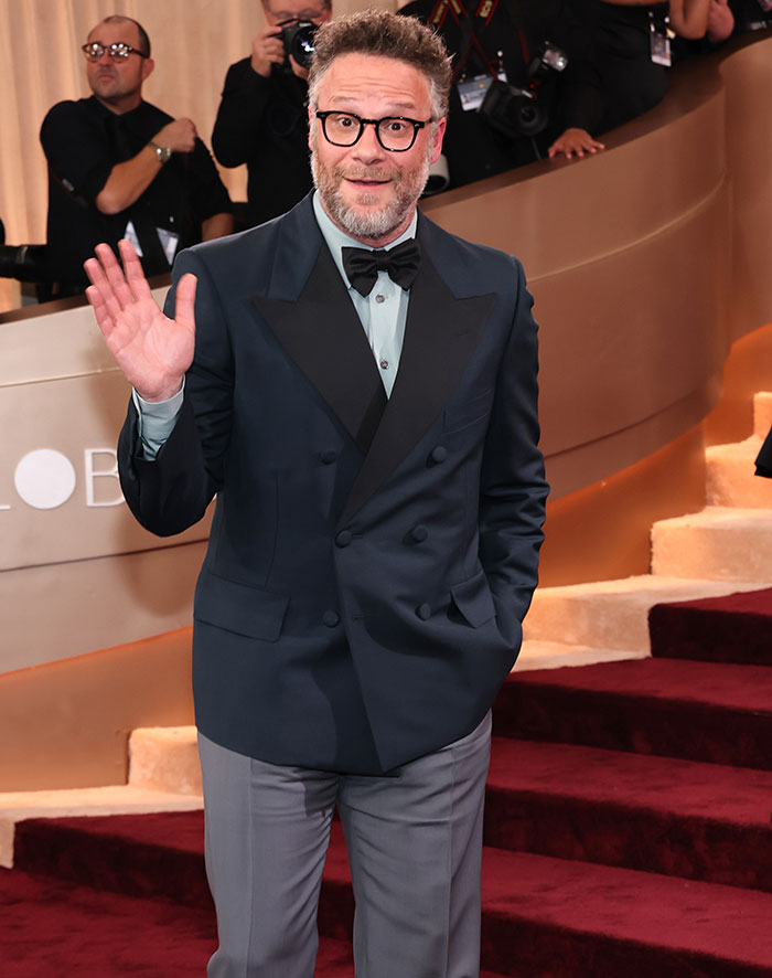 Seth Rogen in a dark suit waving on the red carpet at an awards event amid photographers and media.
