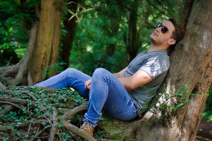Young man wearing sunglasses and casual clothes sitting against a tree in nature, reflecting on crazy party stories nightlife.
