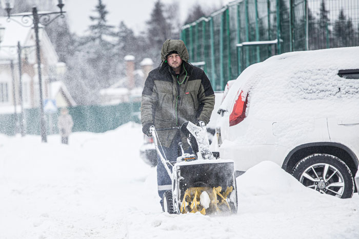 Man clearing snow with a snow blower next to a snow-covered illegally parked car on a residential street. Man clearing snow with a snow blower next to a snow-covered illegally parked car on a residential street.