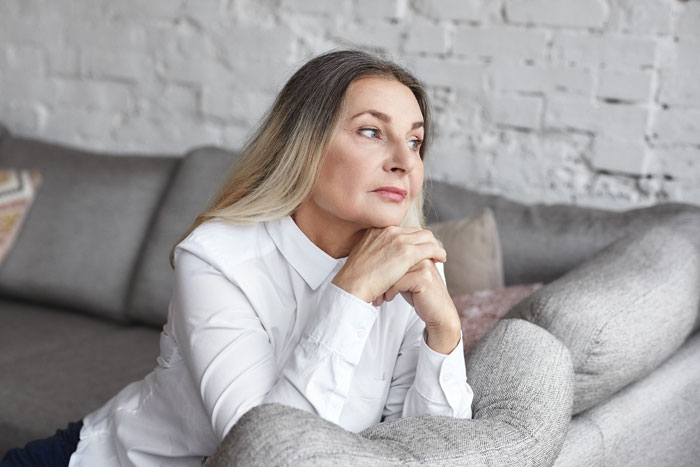 Thoughtful middle-aged woman in white shirt sitting on couch reflecting on son marrying girlfriend after high school advice. Thoughtful middle-aged woman in white shirt sitting on couch reflecting on son marrying girlfriend after high school advice.