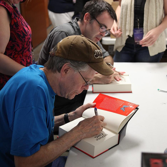 Stephen King wearing a cap and signing a book at a table during a public book signing event.