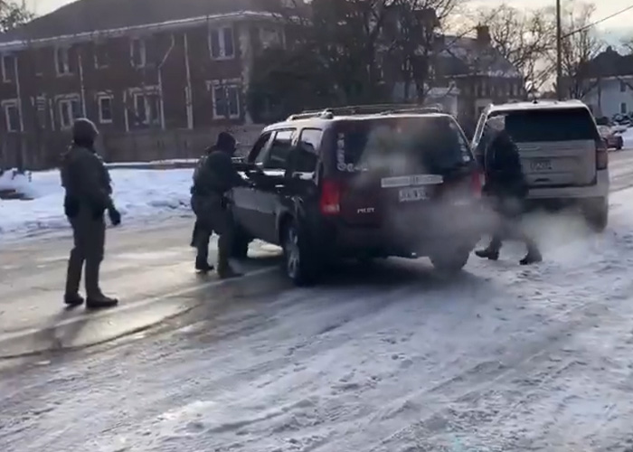 ICE agents in tactical gear conducting a vehicle stop on a snowy residential street during a law enforcement operation.