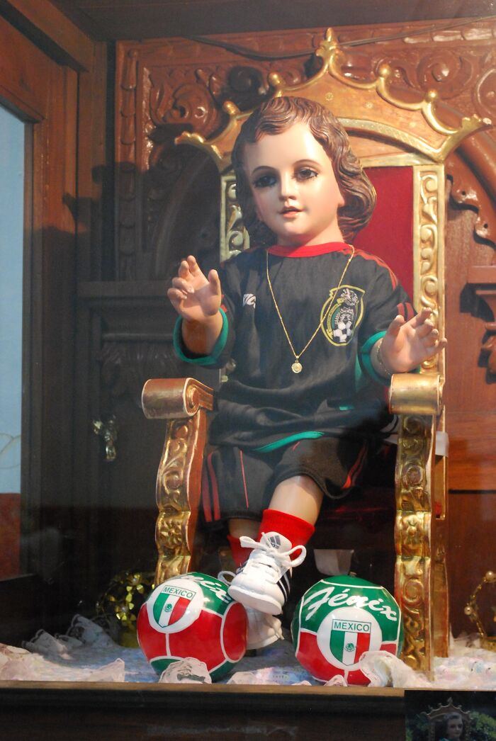Religious tradition statue dressed in soccer gear seated on a golden throne with Mexican soccer balls nearby.