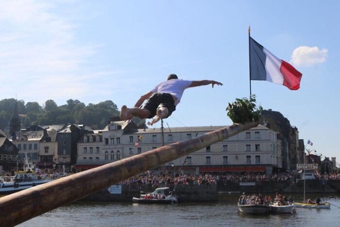 Person leaping off a greased pole over water during a strange tradition in a European town with a French flag visible.