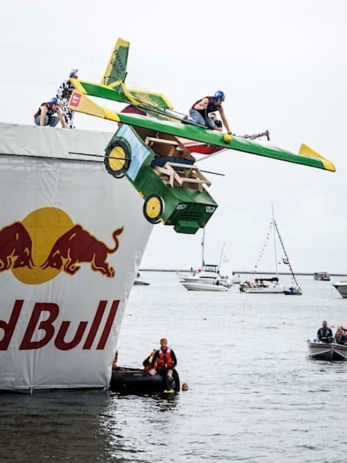 People launching a homemade flying car into water during a strange tradition event with boats in the background.