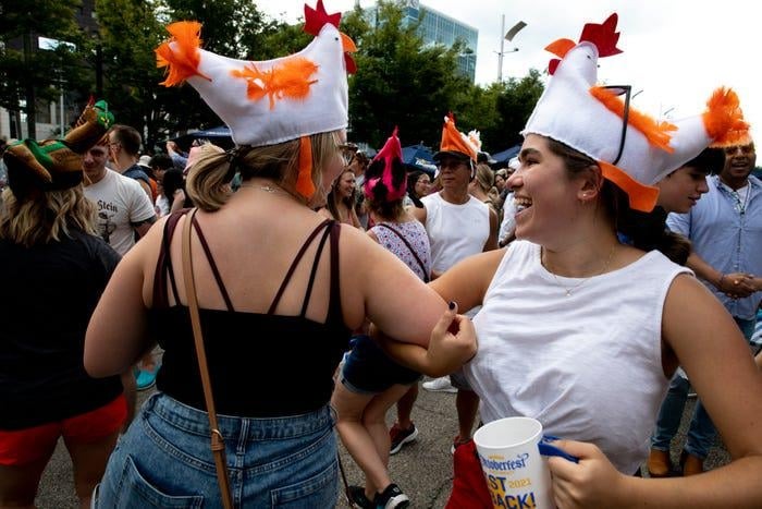 Two women wearing chicken hats laugh and dance outdoors during a strange traditions festival crowd.