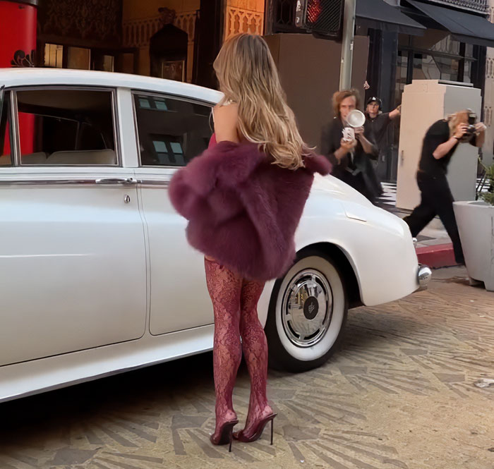 Sydney Sweeney in red lace lingerie and a fur coat poses next to a vintage white car while photographers capture her in LA.