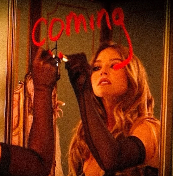 Sydney Sweeney writing on mirror wearing sheer gloves, promoting her new racy lingerie line in a stylish setting.